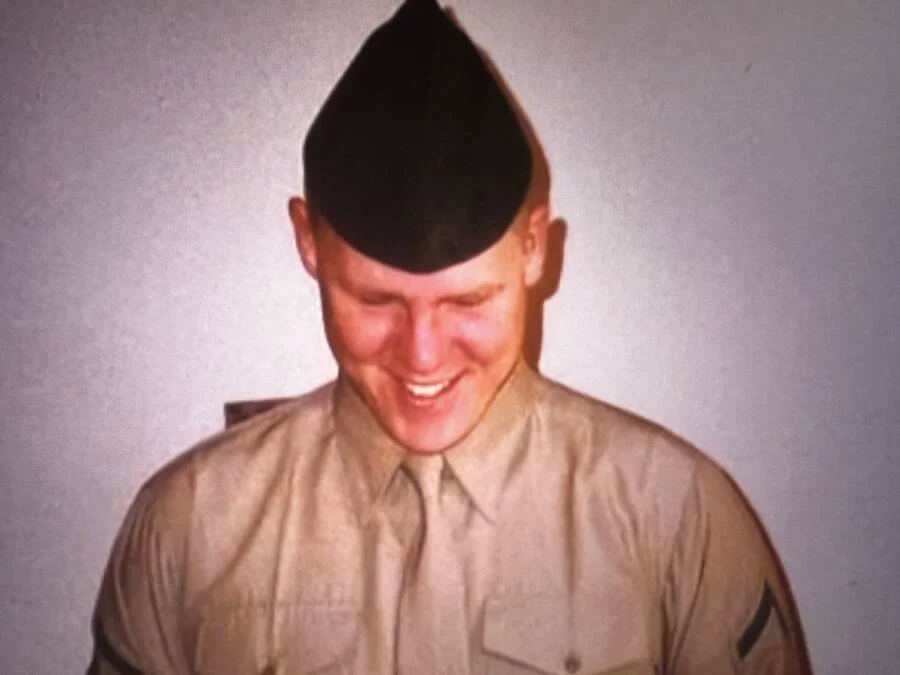 Nathan Gale com uniforme da marinha americana de frente para uma parede branca. Ele sorri olhando para baixo.